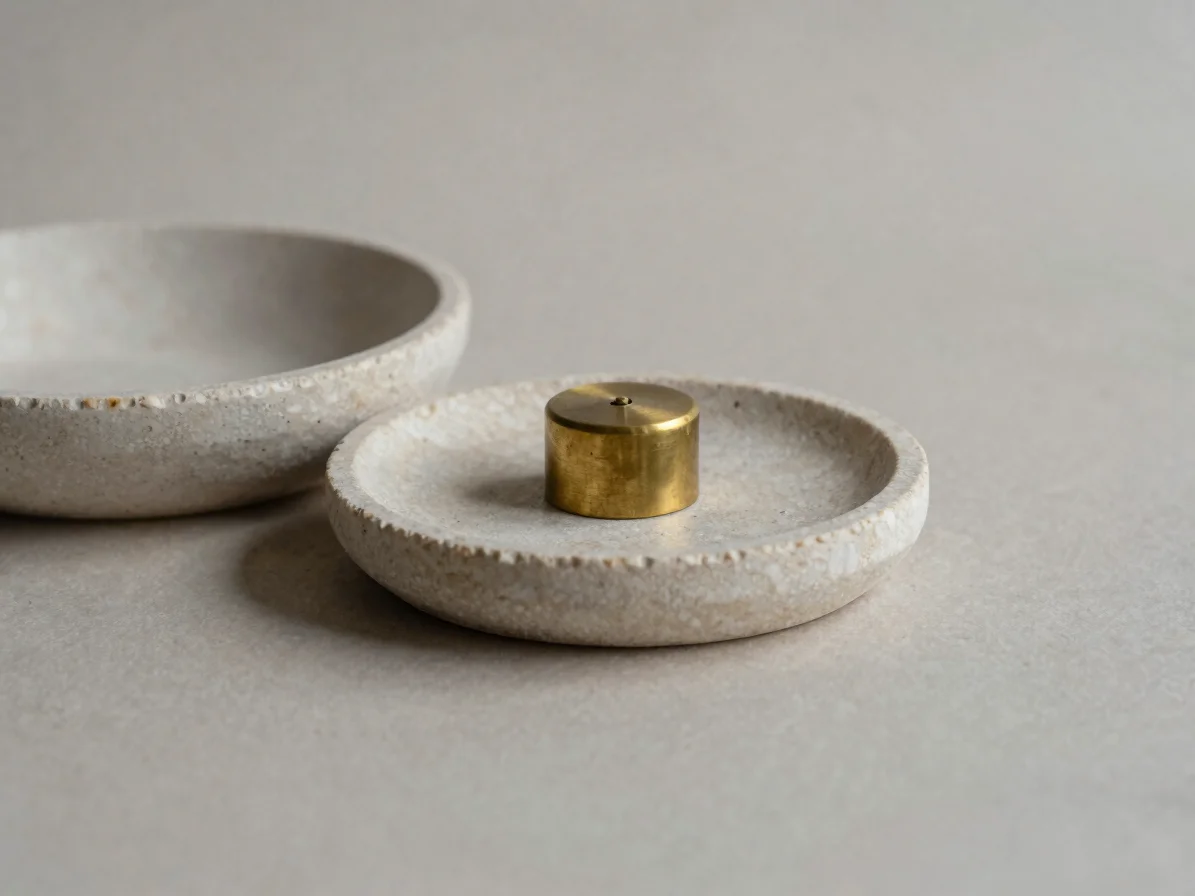 Muted editorial product photograph of a small brass-finished object beside a pale stone dish, soft diffused studio dayli